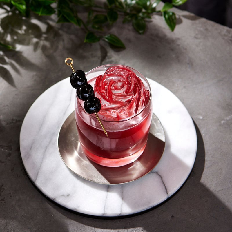 A deep red cocktail with skewered cherries on a marble coaster, foliage hanging into the frame above