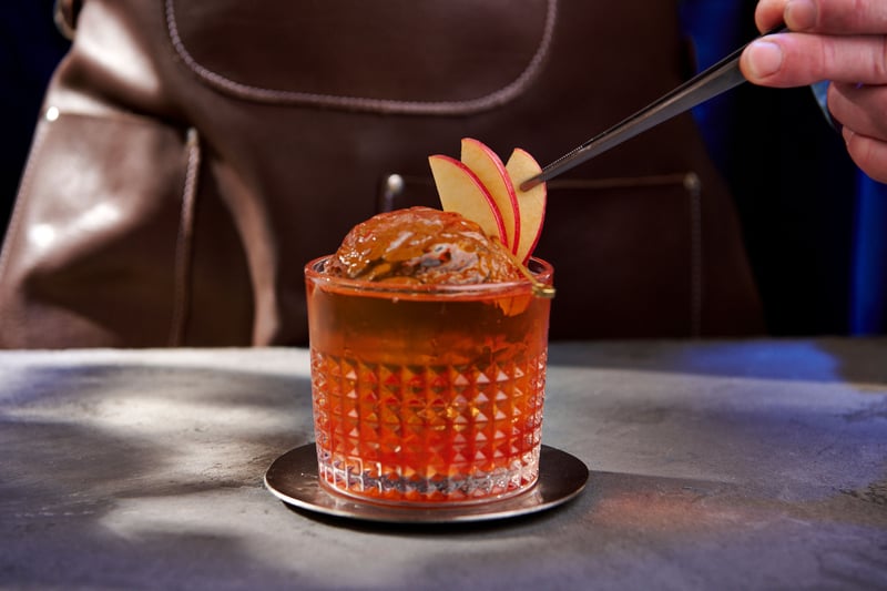 A hand lowers a garnish into an old fashioned on a dark bar, the glass lit from behind