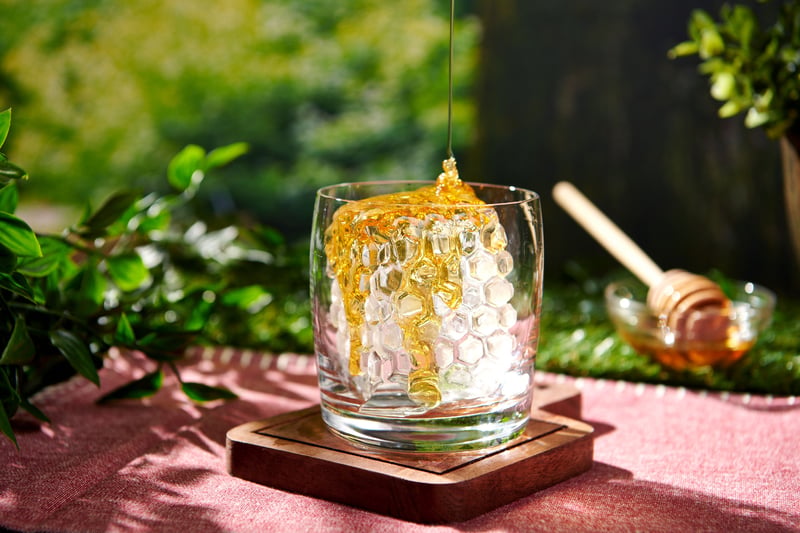 Honey drizzles over a glass of iced drink on a wooden coaster, set on a red cloth with greenery