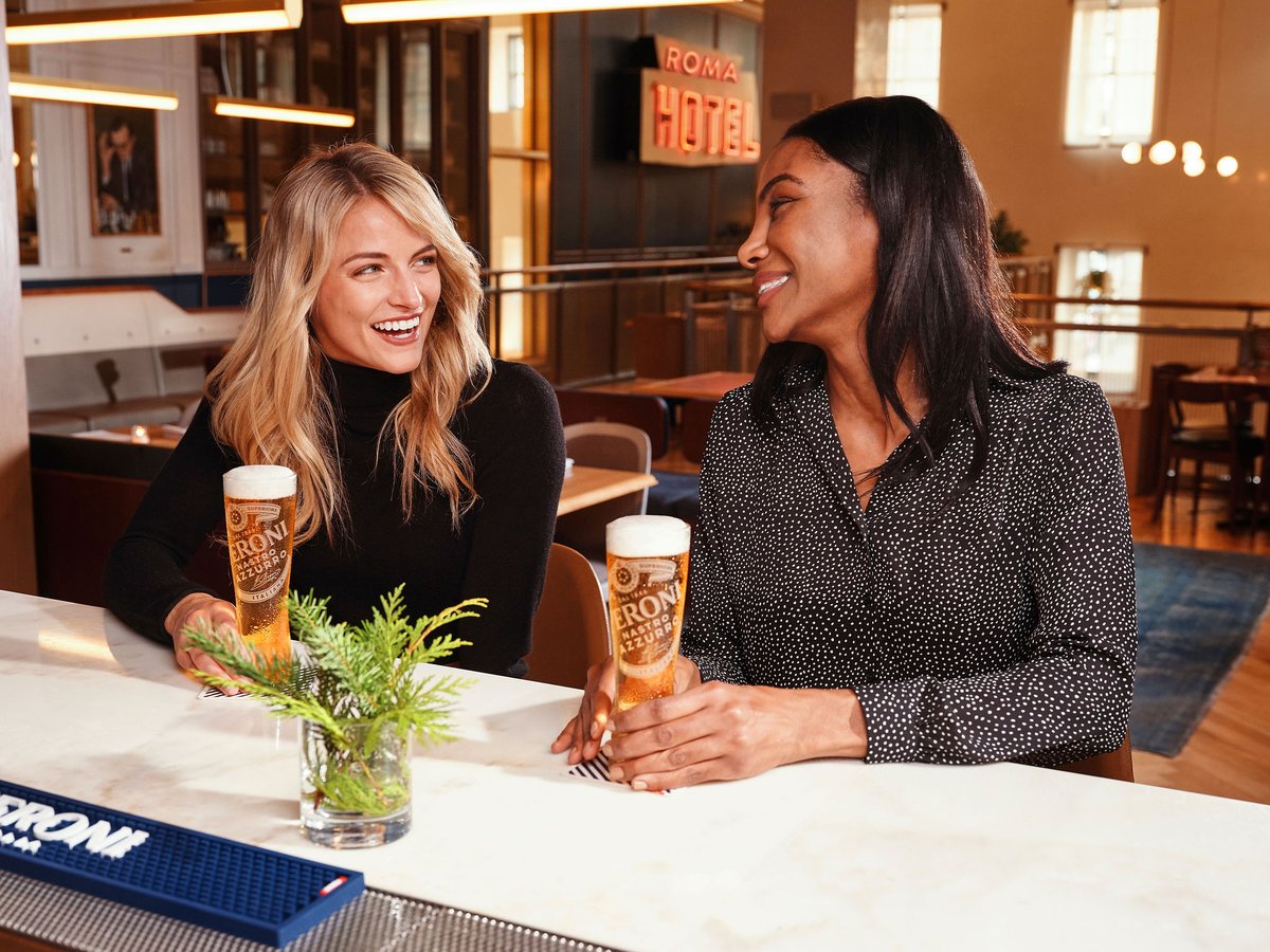 Two women talk over tall glasses of lager at a marble bar counter