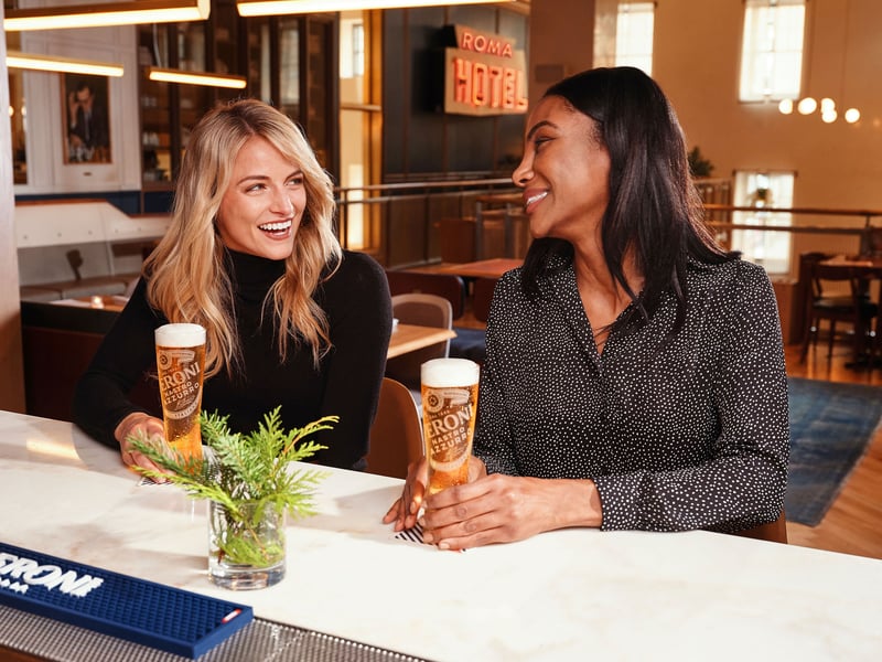 Two women talk over tall glasses of lager at a marble bar counter