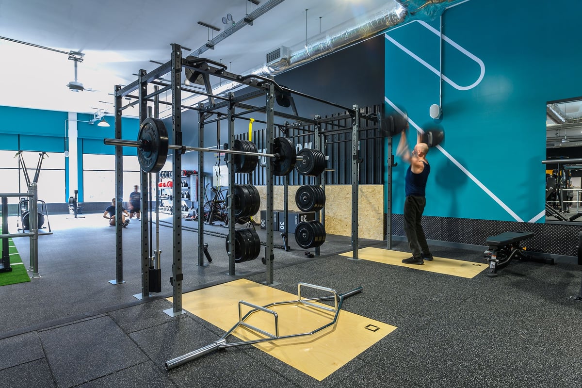 PureGym Bath Spring Wharf: Squat racks, weight plates, rubberized flooring, and mirrors in a gym with teal accent walls.