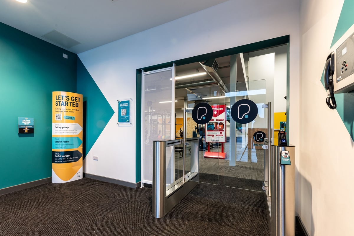 PureGym Heanor: Vestibule with self-service kiosks, glass doors, teal walls, signage.