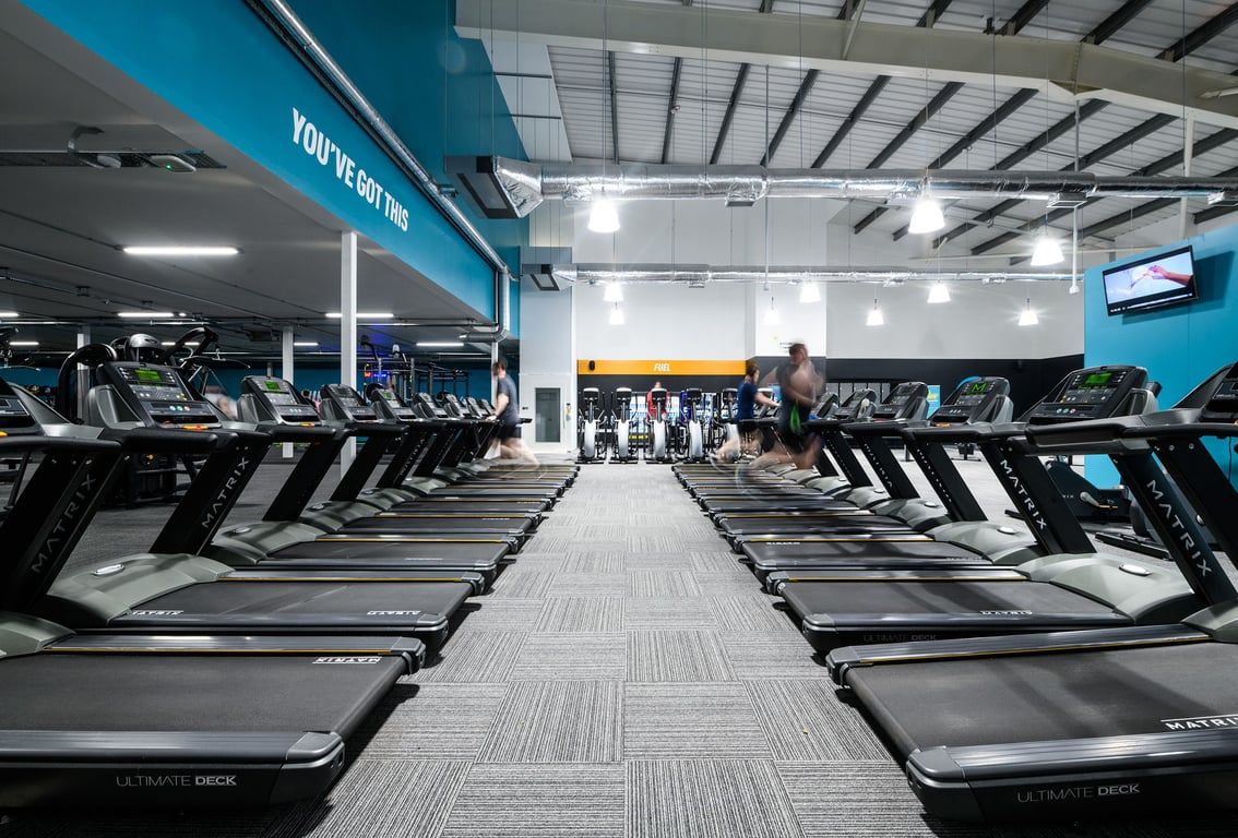 PureGym Cardiff Bay: Rows of treadmills arranged in an indoor gym space with teal accents and ample lighting.