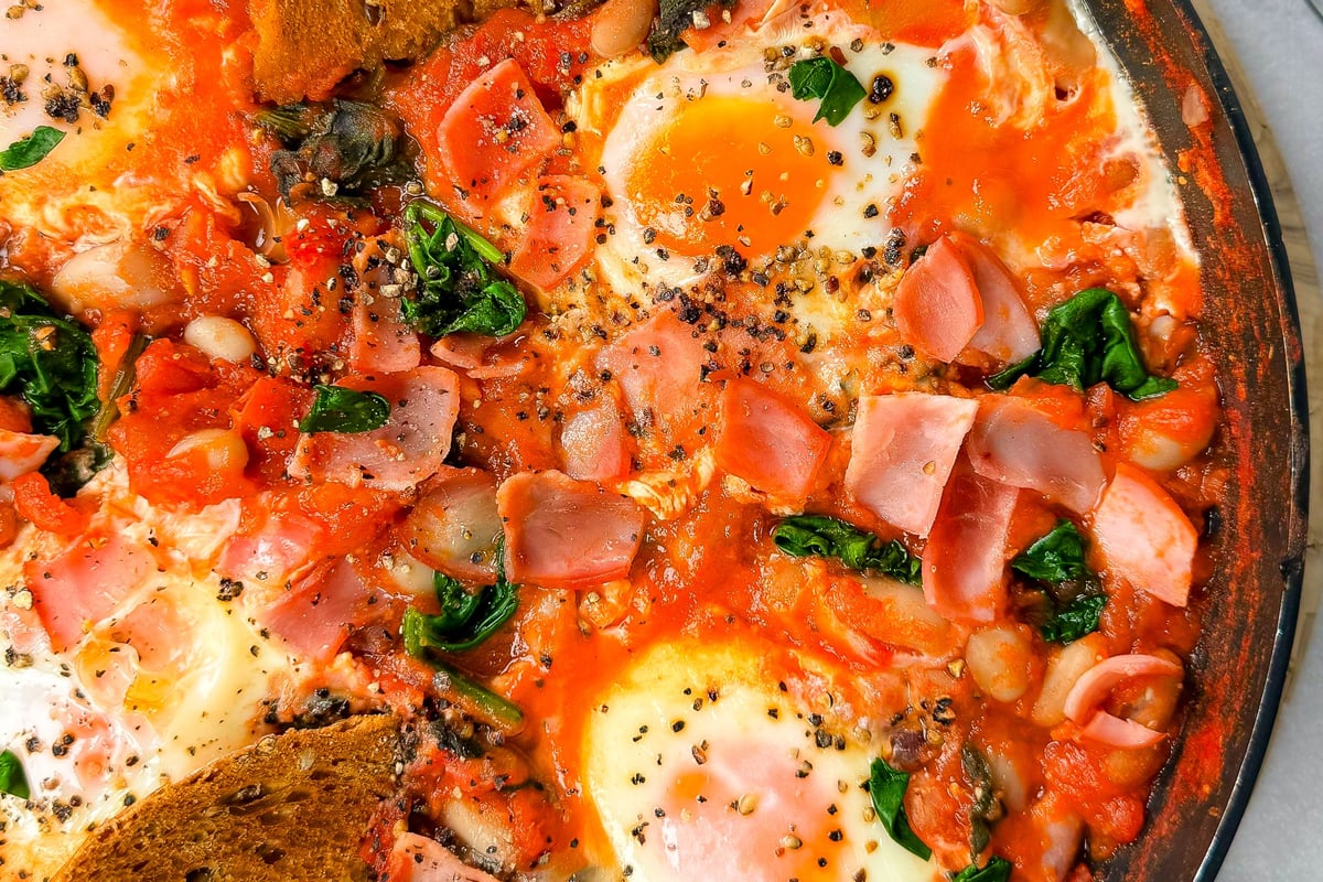 Shakshuka with eggs, spinach, ham, beans, tomato sauce, and toasted bread slices in pan