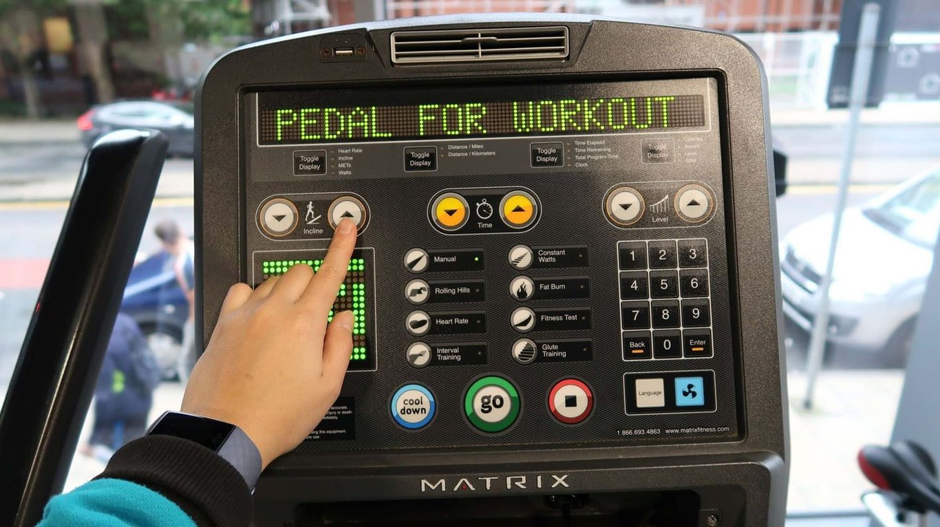 Hand pressing incline button on exercise bike console with PEDAL FOR WORKOUT text above
