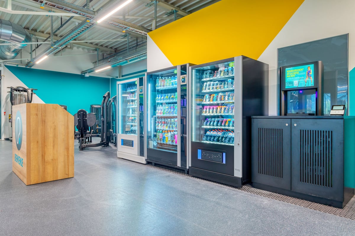 PureGym Martigny: Kühlschränke mit Getränken, braunen Boxen und schwarzen Verkaufsautomaten.