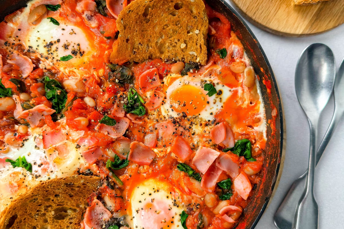 Shakshuka with eggs, spinach, ham, and toasted bread in a pan with two spoons nearby