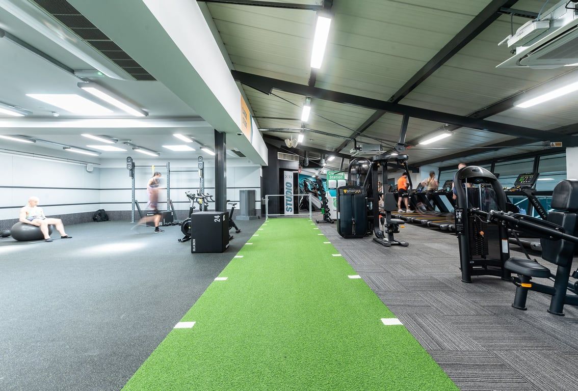 PureGym London Bayswater: Weights, machines, green turf area for training, fans on ceiling in a well-equipped gym interior.