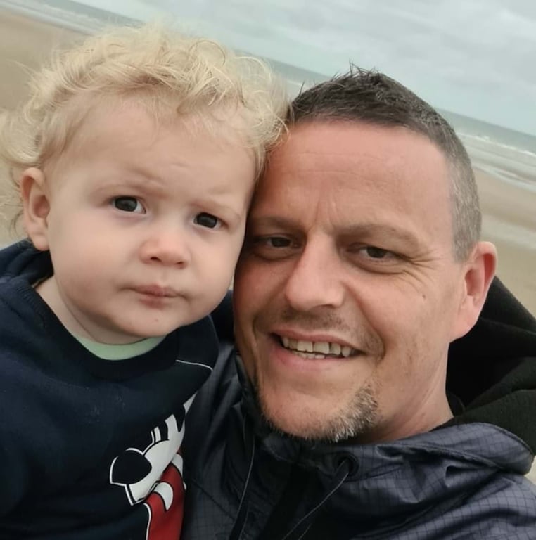 Man smiling holding a toddler with curly blonde hair on a beach background