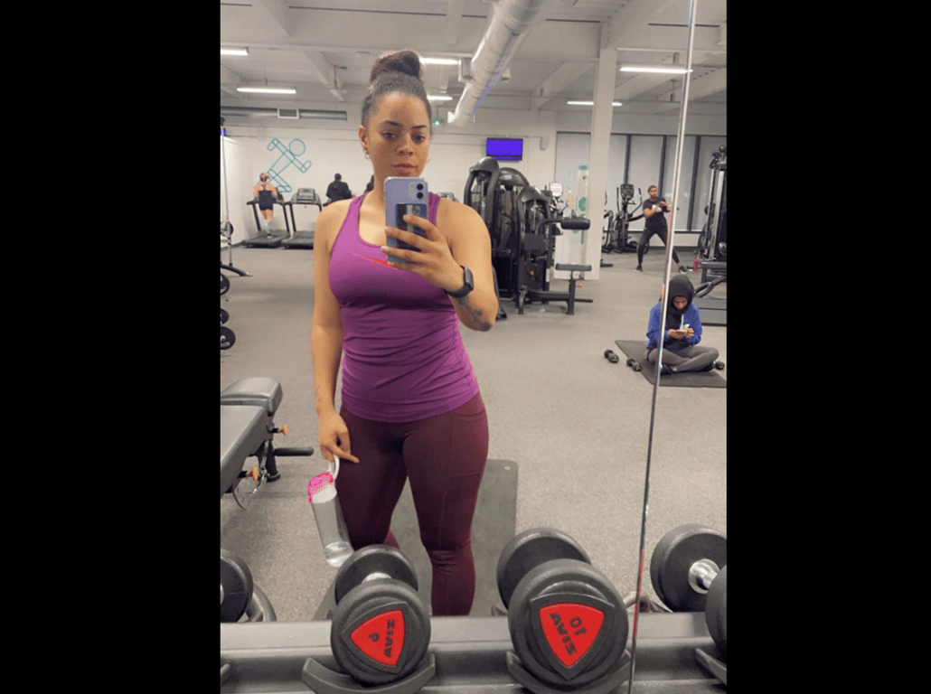 Woman in purple workout clothes taking a mirror selfie in a gym with dumbbells in front