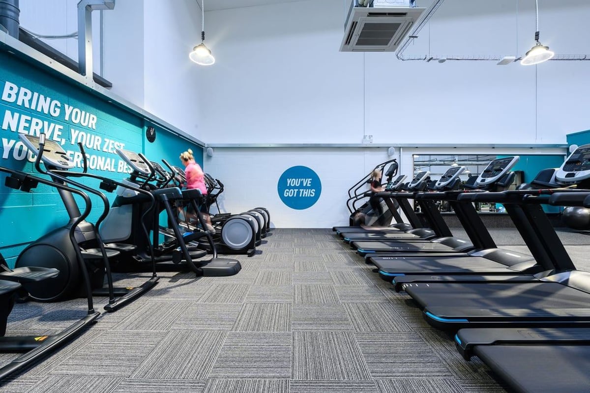 PureGym Saffron Walden: Rows of upright exercise bikes and treadmills in a gym studio with teal accents.