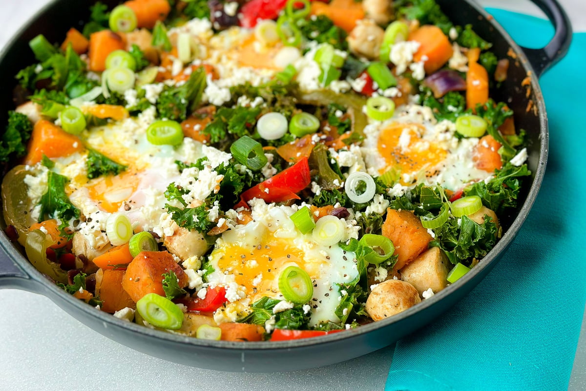 Skillet of sweet potato hash with fried eggs, kale, peppers, spring onions and feta