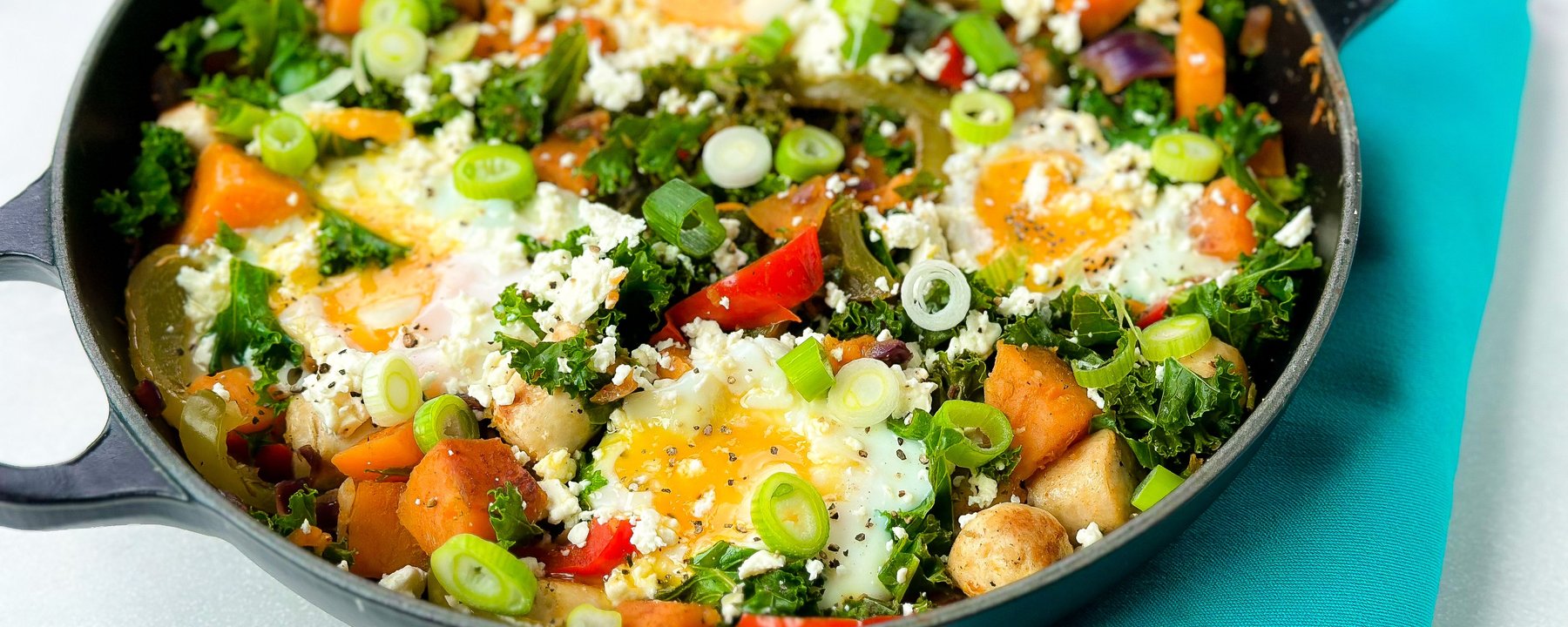Skillet of sweet potato hash with fried eggs, kale, peppers, spring onions and feta