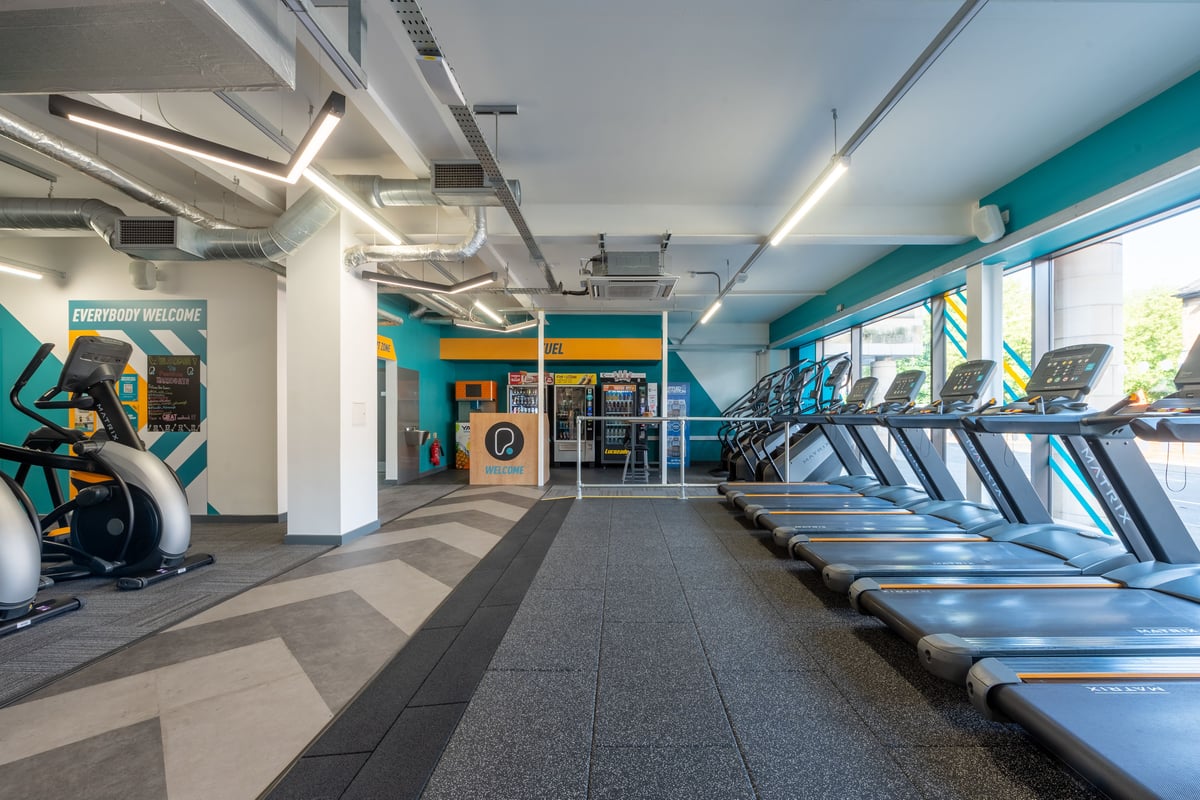 PureGym Harrogate: Rows of treadmills, wall mural artwork, self-service kiosks in brightly lit gym space.