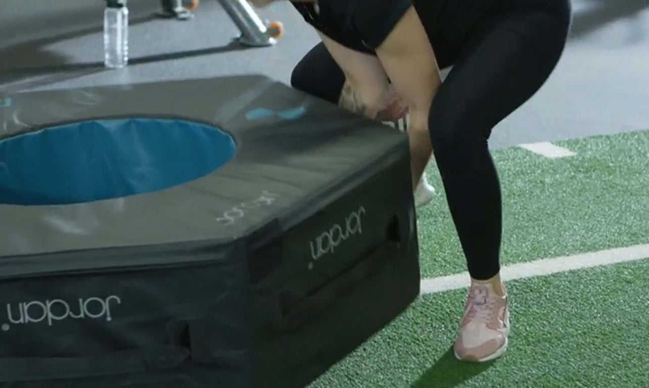 Person in black leggings and pink trainers lifting a padded exercise box on turf flooring