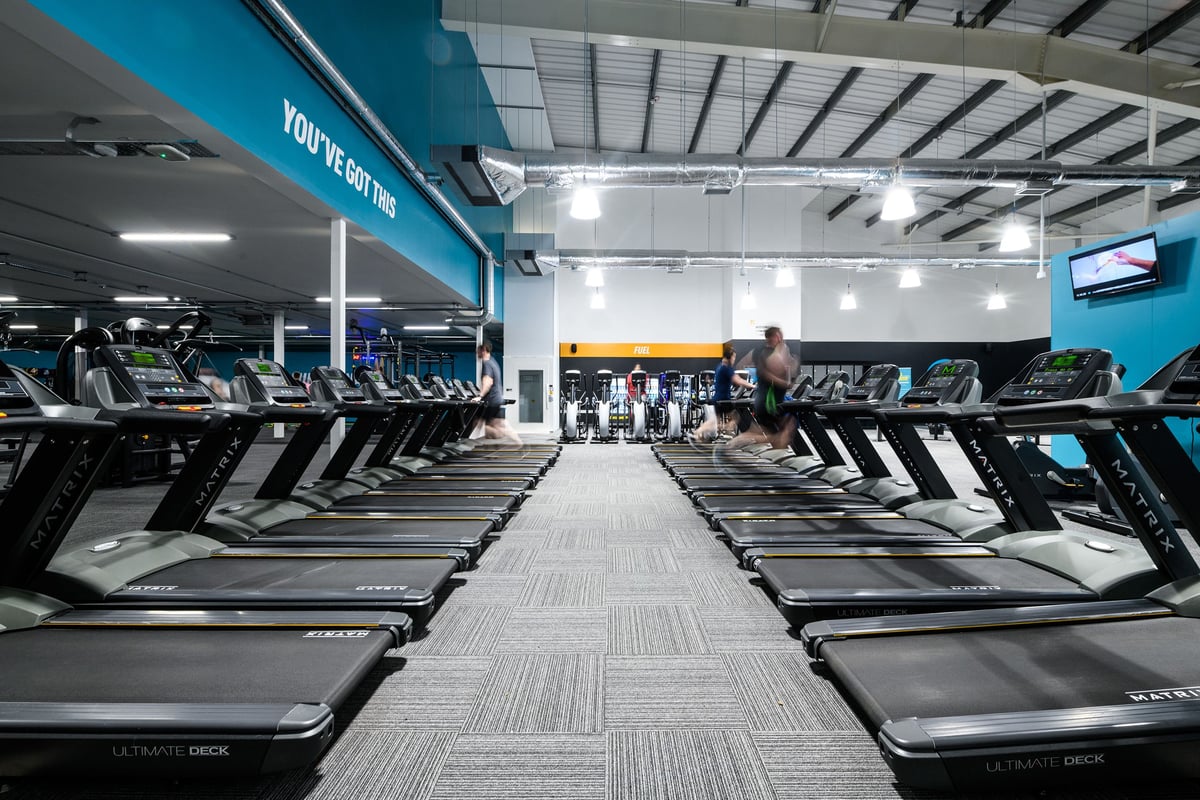 PureGym Cardiff Bay: Rows of treadmills arranged in an indoor gym space with teal accents and ample lighting.