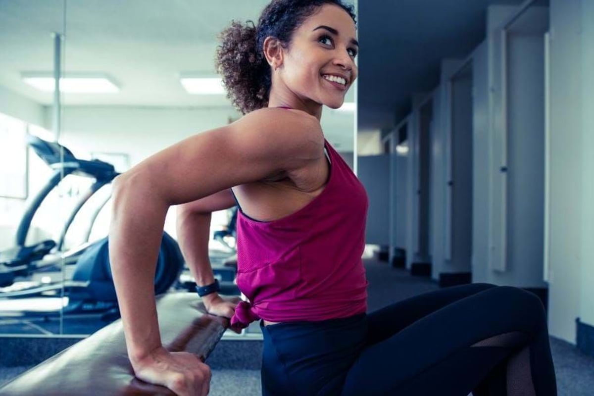 Woman in purple top doing tricep dips on bench in gym with cardio machines.