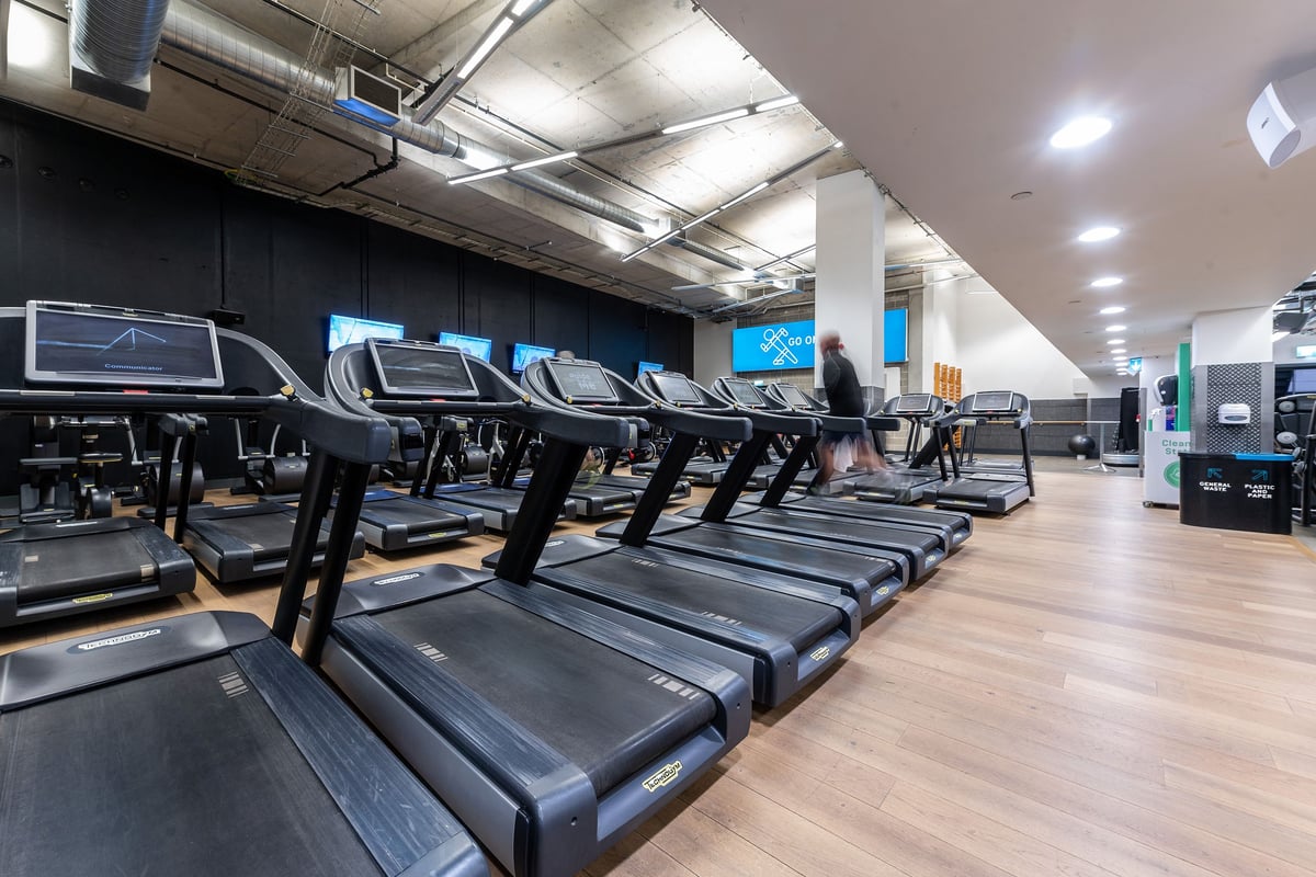 PureGym London Aldgate: Rows of treadmills inside a dimly lit gym space with industrial ceiling pipes.