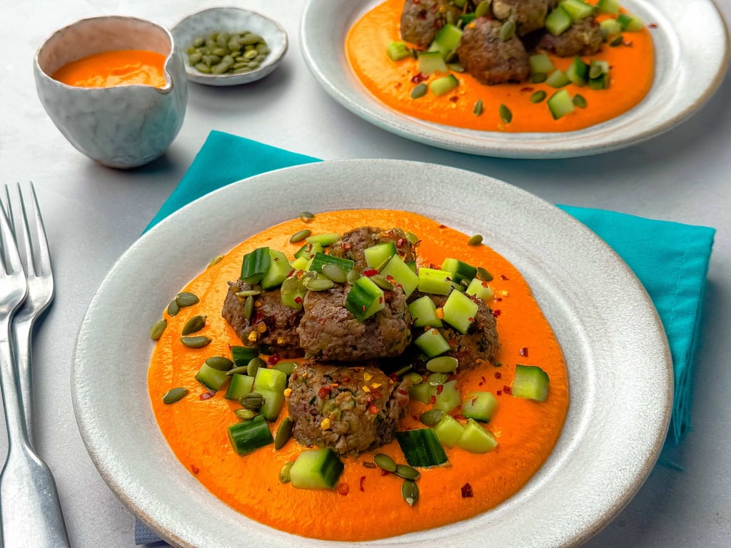 Meatballs on orange sauce topped with diced cucumber and pumpkin seeds on white plates.