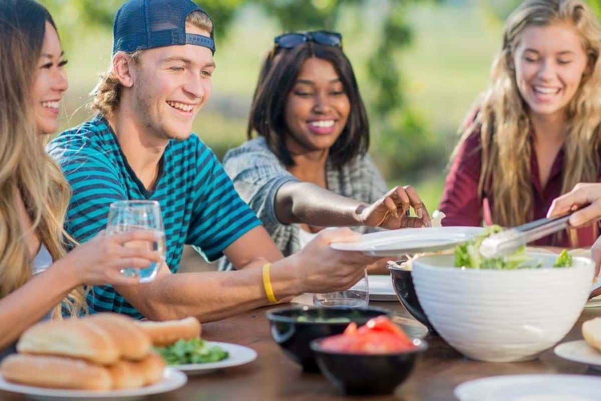 Group of young people smiling and serving salad at outdoor table meal