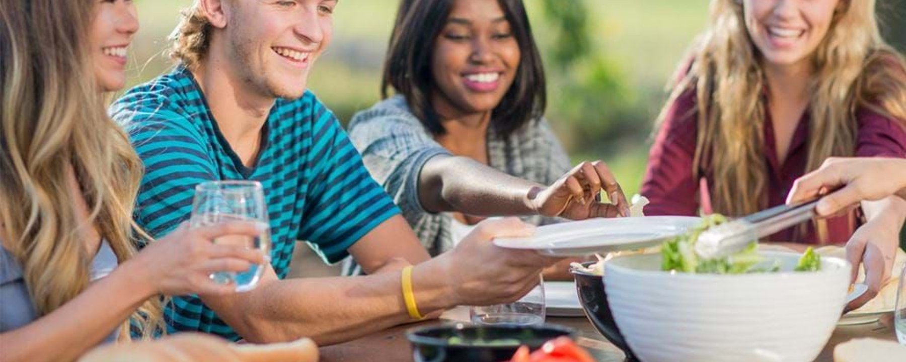 Group of young people smiling and serving salad at outdoor table meal