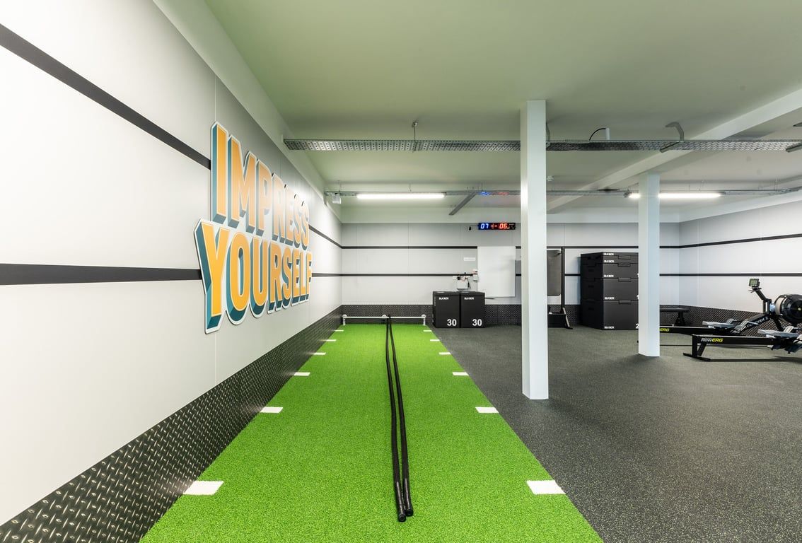 PureGym Glasgow Giffnock: Green turf, weights area, ceiling-mounted rigs in a basement-style gym interior.