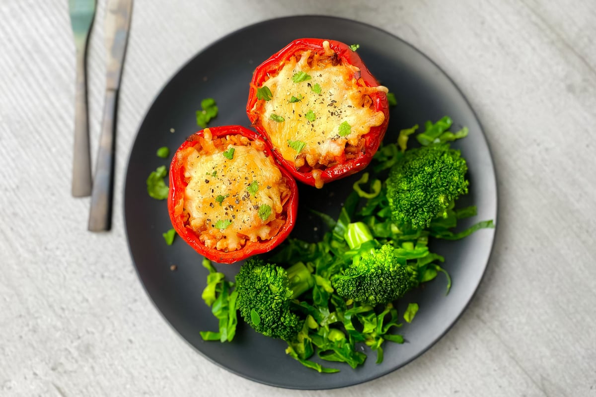 Two stuffed red peppers with melted cheese topping served with broccoli on a black plate