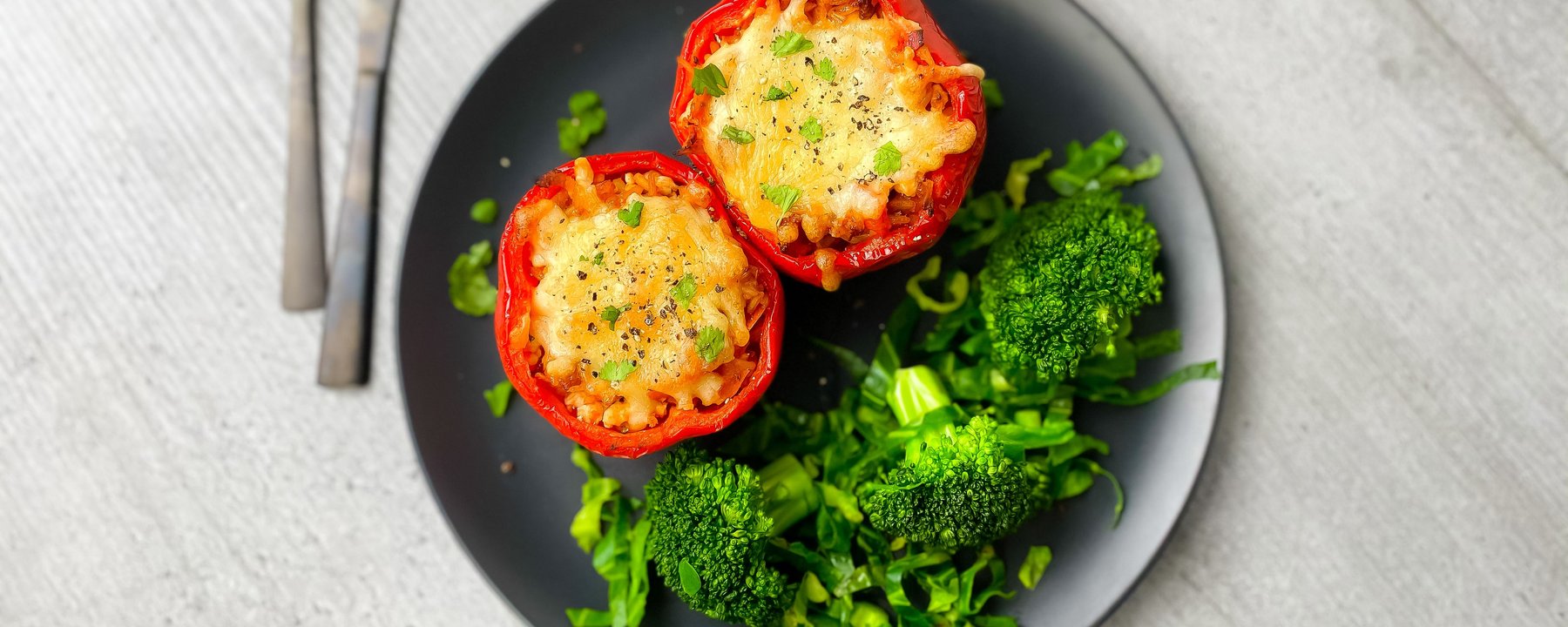 Two stuffed red peppers with melted cheese topping served with broccoli on a black plate