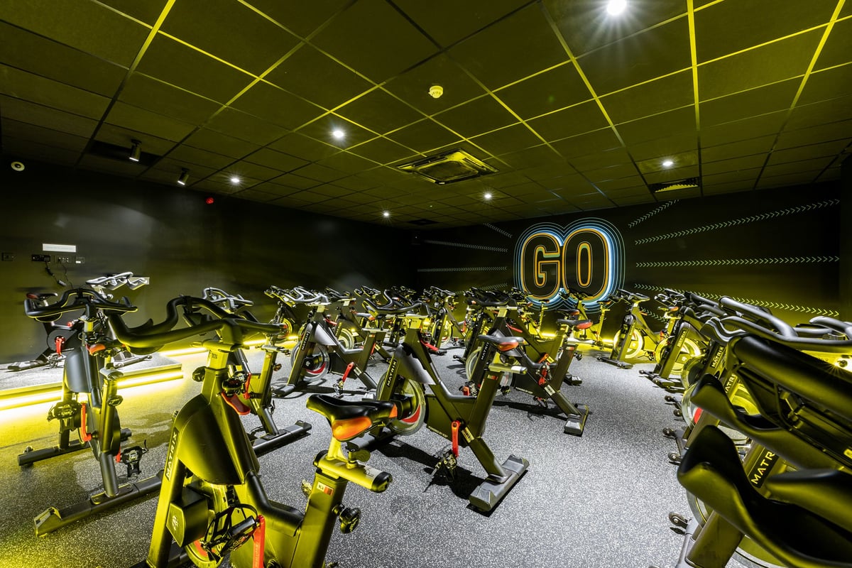 PureGym Gloucester Retail Park: Rows of stationary cycles in a dimly lit room with yellow lighting and "GO" signage.
