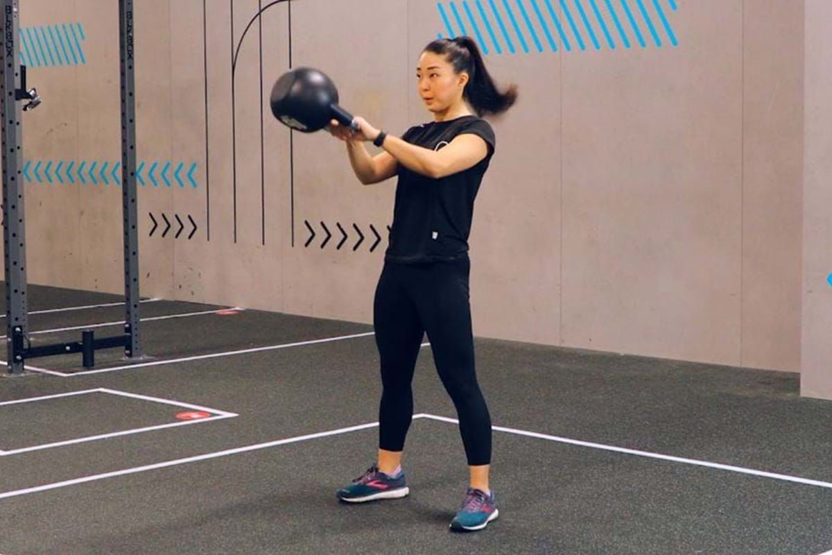 Woman in black workout clothes swinging a kettlebell in a gym with grey walls.