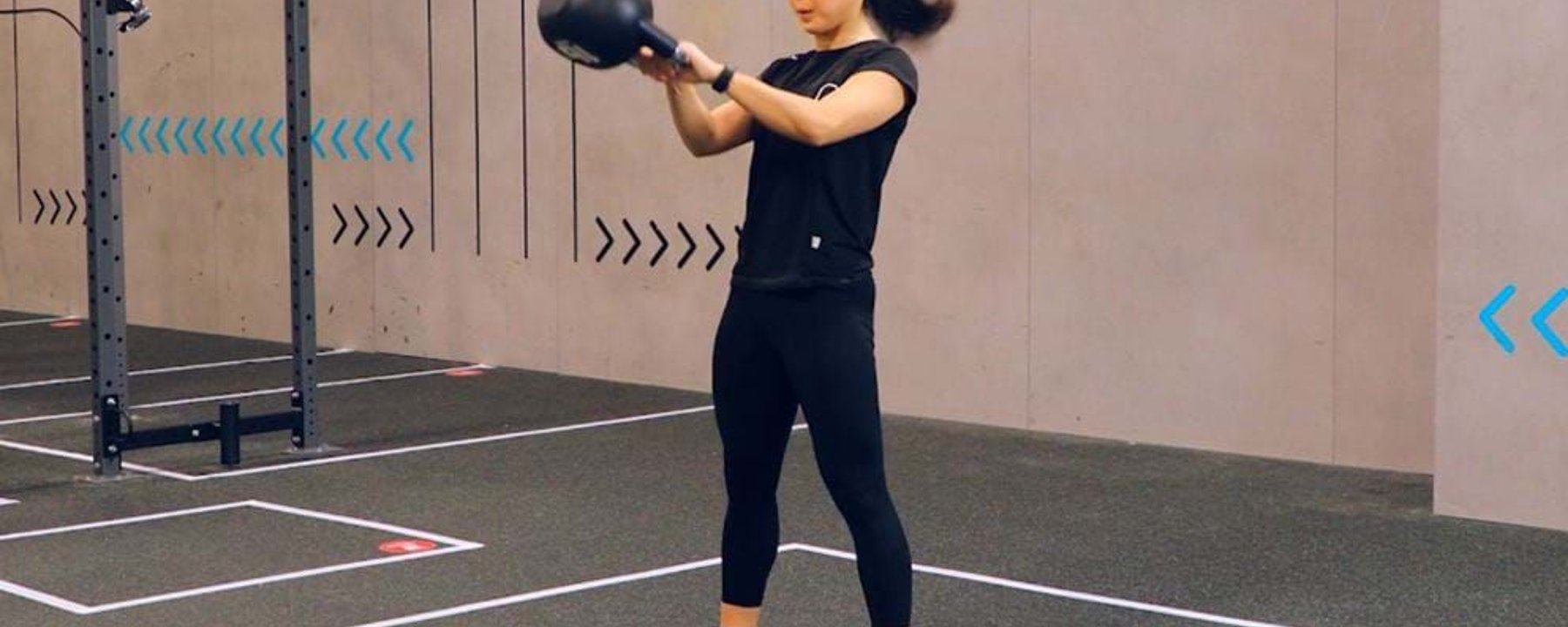 Woman in black workout clothes swinging a kettlebell in a gym with grey walls.