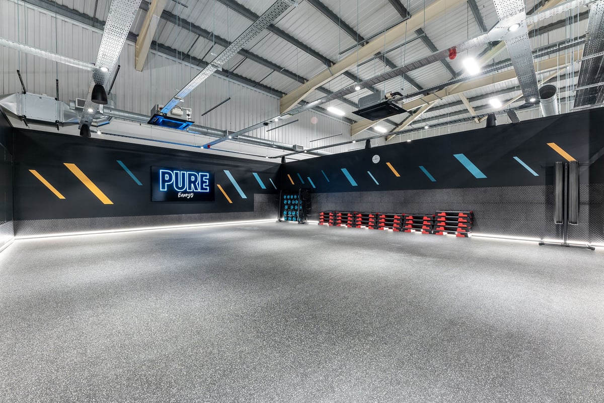 PureGym Merthyr Tydfil: Bright studio with rubberized flooring, neon lighting, weight plates, and resistance machines.