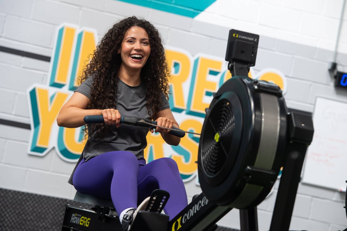 Woman with curly hair smiling while using a rowing machine in a gym