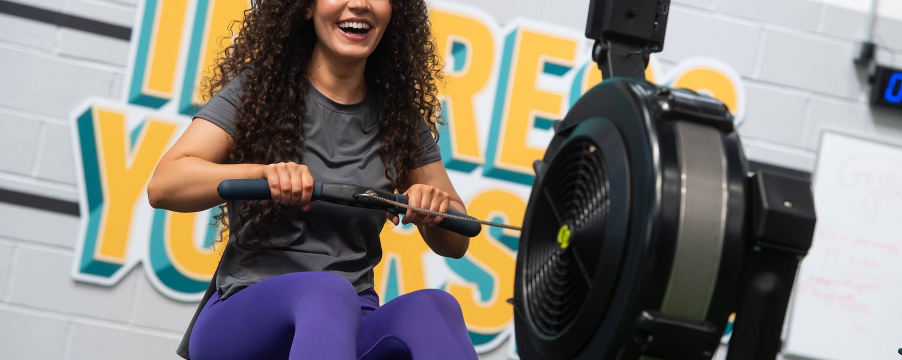 Woman with curly hair smiling while using a rowing machine in a gym