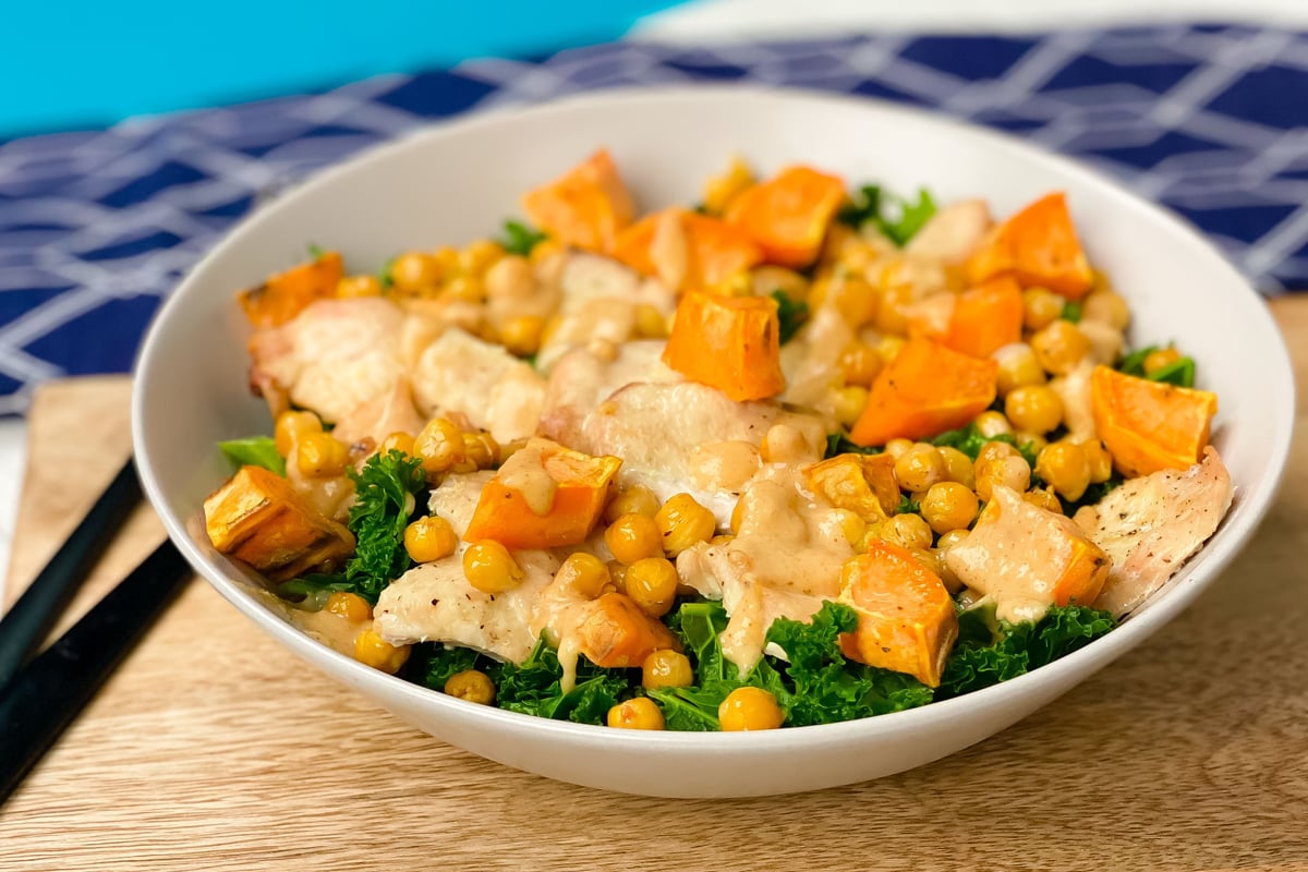 White bowl containing sliced chicken with chickpeas, sweet potato and kale on wooden board