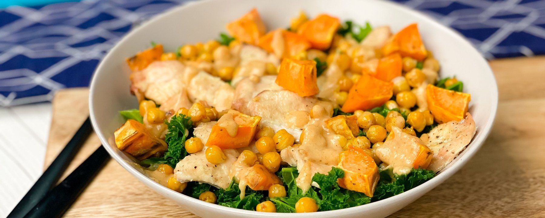White bowl containing sliced chicken with chickpeas, sweet potato and kale on wooden board