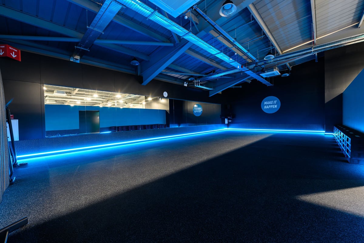 PureGym St Albans: Blue-lit empty studio space with rubberized flooring and illuminated support beams.