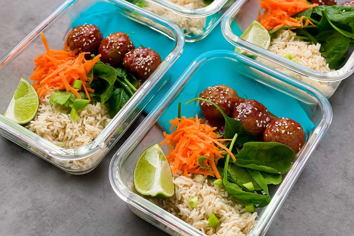 Meal prep containers with meatballs, rice, shredded carrot, spinach and lime wedges