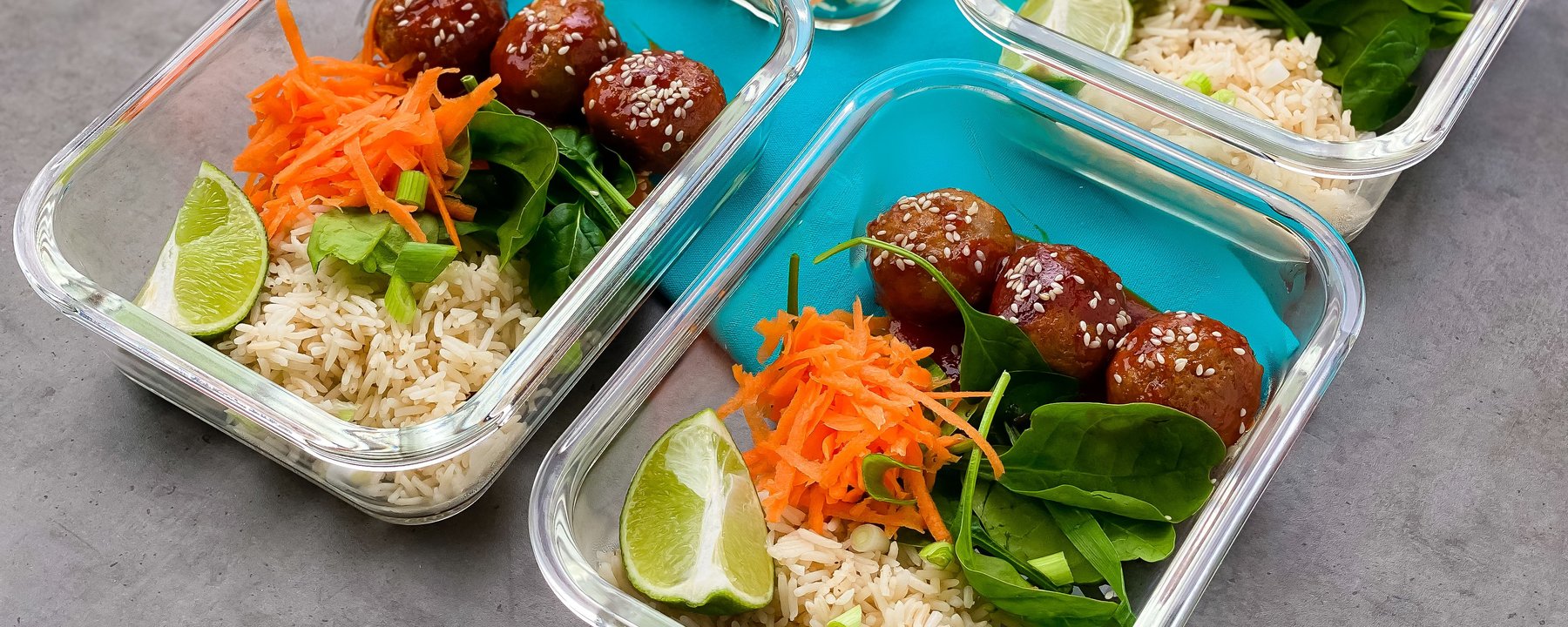 Meal prep containers with meatballs, rice, shredded carrot, spinach and lime wedges