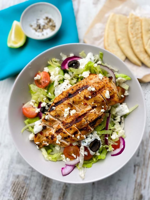 Grilled chicken breast on a salad with lettuce, tomatoes, olives, onions, and feta cheese.
