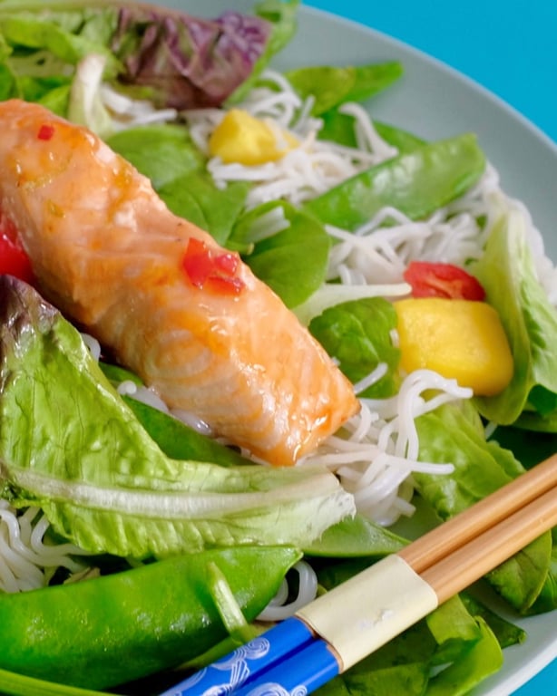 Grilled salmon on a bed of rice noodles, leafy greens, mango, and chopsticks on a plate