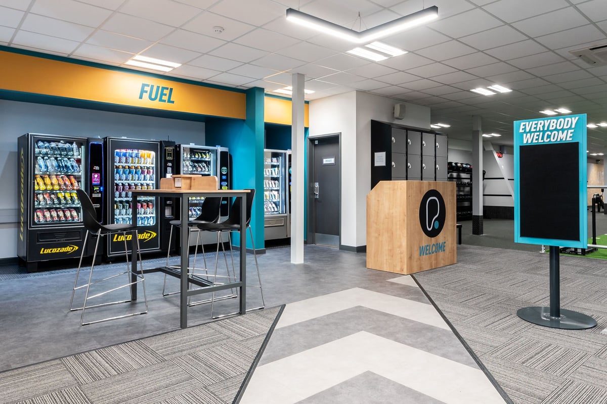 PureGym Gloucester Retail Park: Vending machines with snacks and drinks, a wooden kiosk, and seating area.
