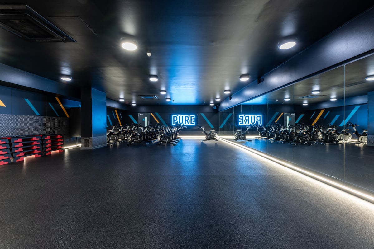 PureGym London Edgware: Rows of stationary bikes facing mirrored walls in a dimly lit studio with blue lighting.