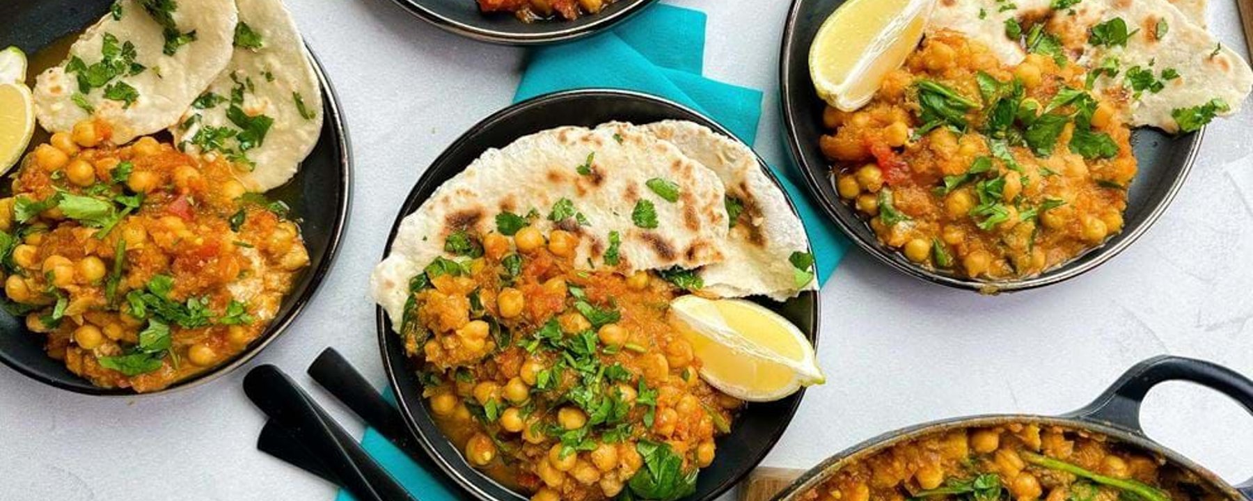 Bowls of chickpea curry with flatbread and lemon wedges on a white surface