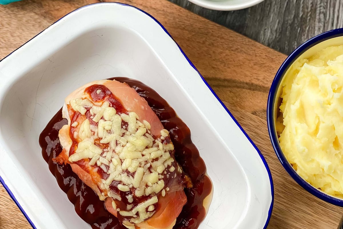 Chicken breast topped with cheese and sauce in a white dish with green beans and mash.