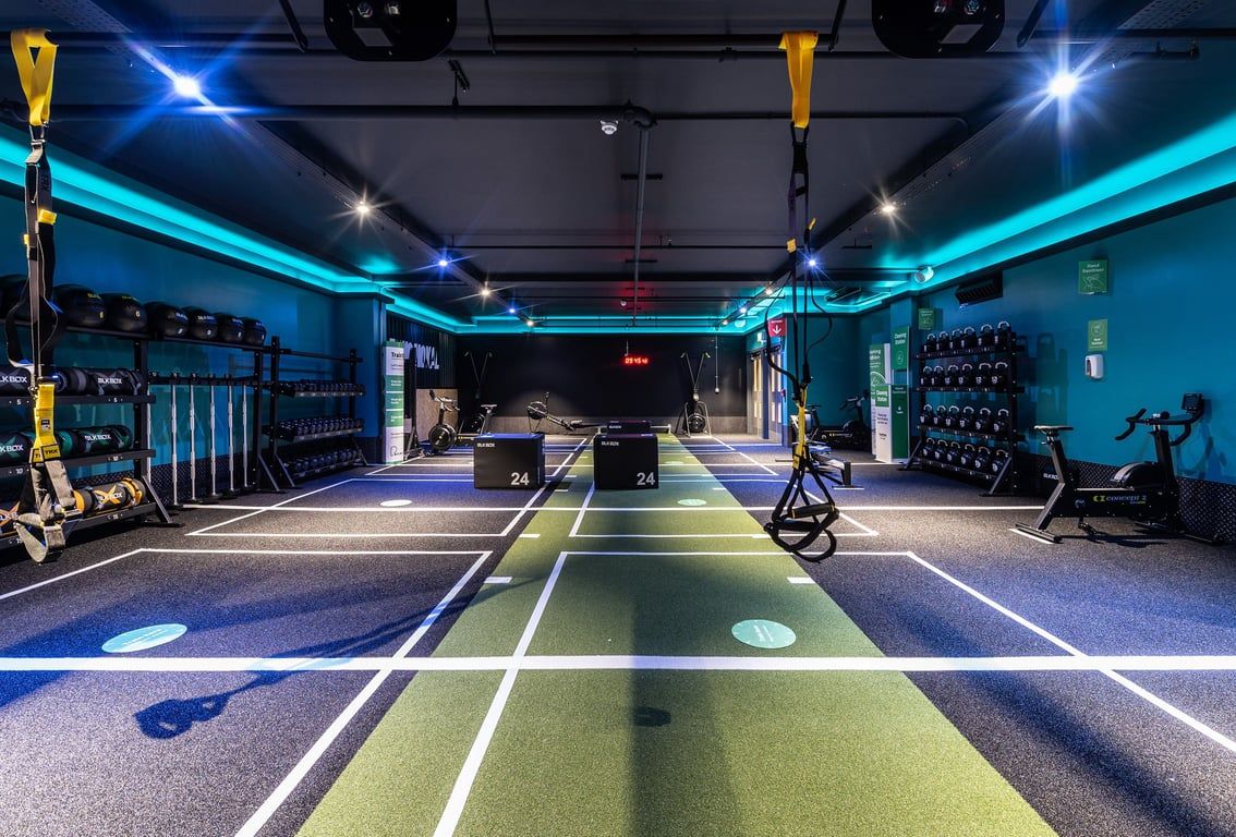PureGym London Bank: Squat racks, benches, free weights, rubberized flooring in a dimly lit studio with blue accent lighting.