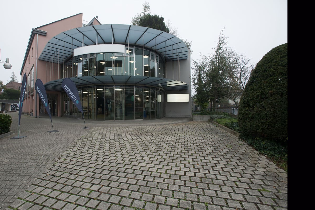 PureGym Lyss: Extérieur du bâtiment moderne en verre et en brique, cour pavée devant l'entrée en verre incurvé.