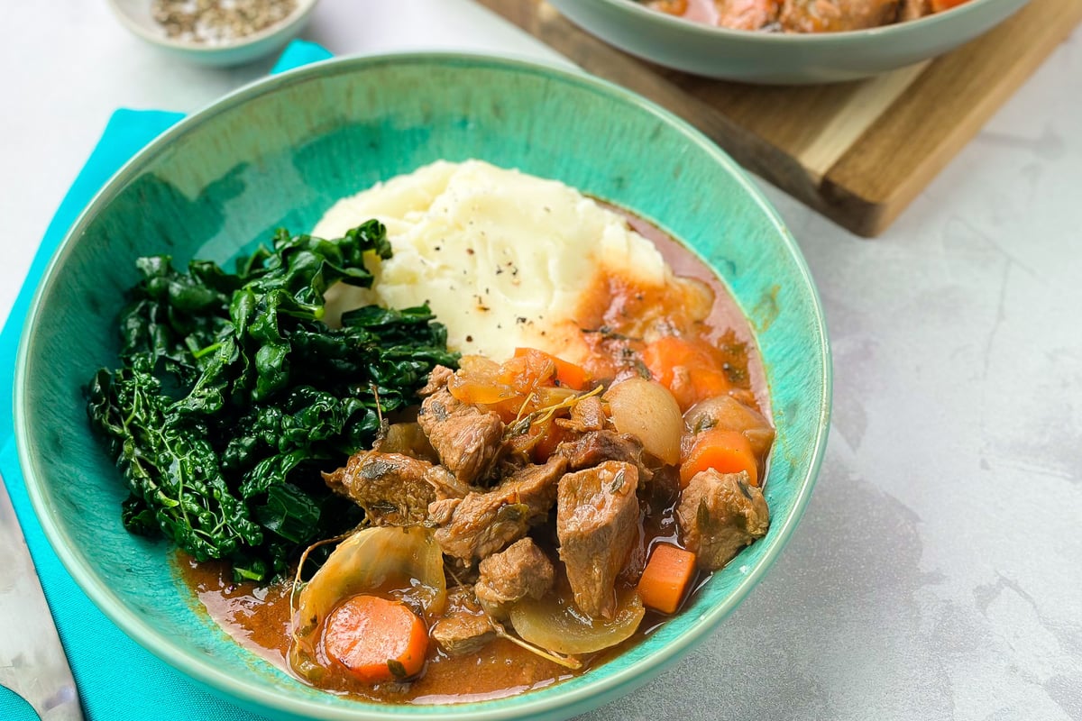 Beef stew with carrots and onions served in turquoise bowl with mashed potato and greens