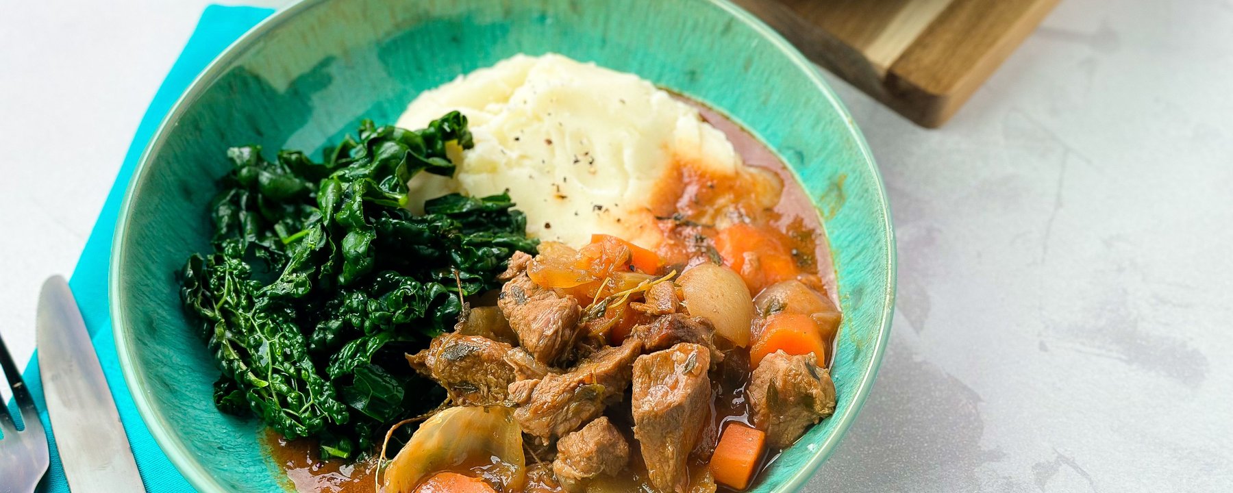 Beef stew with carrots and onions served in turquoise bowl with mashed potato and greens